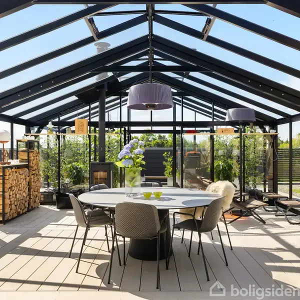 Et glashus med et rundt bord og stole centralt. Træstabel og en pejs til venstre. Haven omgiver strukturen, mens hængende lamper lyser op ovenfra.