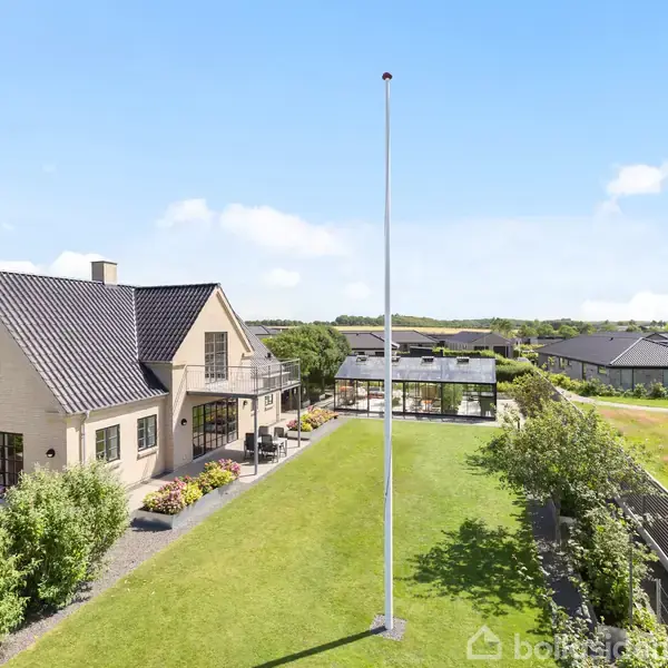 Hus med sort tag og lyse ydervægge står i en velplejet have med flagstang. Omringet af grønne buske og moderne huse, på en solrig dag under blå himmel.