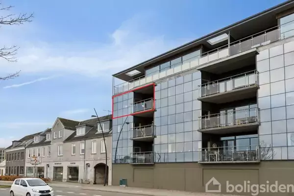 En moderne lejlighedsbygning med glasfacade viser en markeret balkon. I forgrunden kører en hvid bil. Bygningen ligger ved en vej med andre mindre bygninger langs siden.