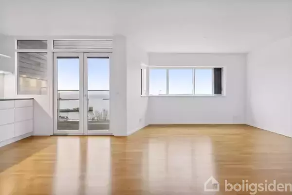 Lys stue med trægulv, store vinduer langs væggen og en dobbelt glasdør til venstre, der åbner til en balkon med udsigt. Minimalistisk interiør uden møbler.