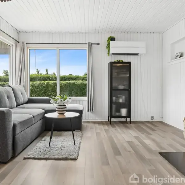 Stue med grå sofa ved vindue, bord med plante foran, og brændeovn til højre. Trægulve og hvid træpanelbeklædt vægge. Hylde med flasker og glasvitrine. Flot udsigt til have.