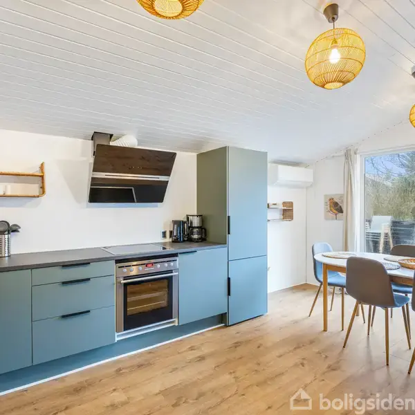 Moderne køkken med blågrønne skabe, integreret ovn og emhætte; tilhørende spiseplads med runde pendellamper og store glasdøre ud til terrasse. Trægulv og hvidmalet loft skaber lys atmosfære.