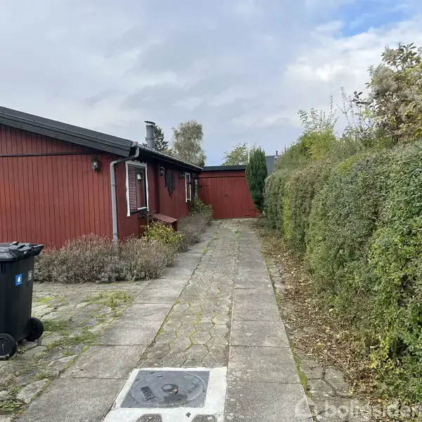 Rødt træhus står stille ved siden af en flisebelagt sti. Til venstre ses affaldsbeholdere, mens en hæk løber langs højre side. Himlen er overskyet.