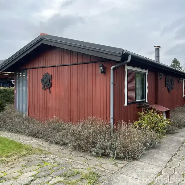Rødt træhus står stille i en have. Det er dekoreret med skodder og emblemer på væggene, og en stenbelagt sti fører forbi. Huset har et mørkt tag.