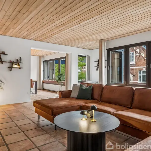 Brun lædersofa står i en stue med flisegulv og træloft. Foran sofaen er et rundt bord. Store vinduer viser en udendørs murstensbygning. En plante står ved siden af en væglampe.