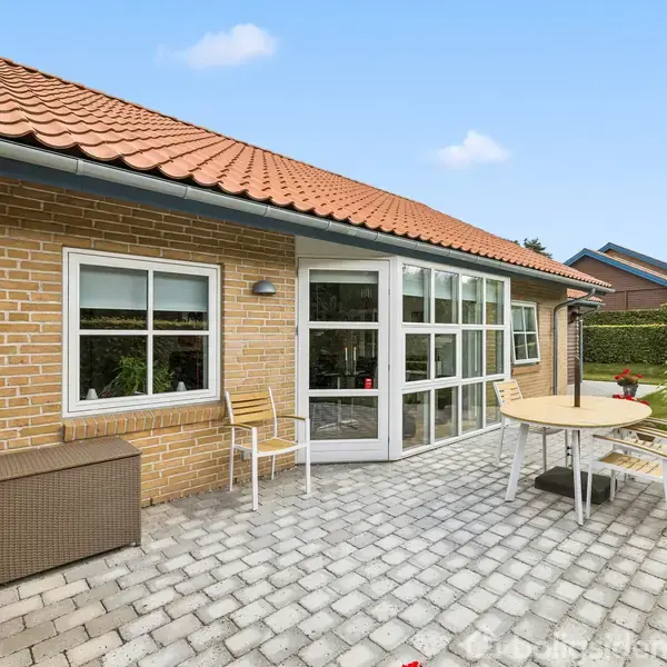 Et stenterrasseområde viser et sæt havemøbler med et bord og flere stole. Terrassen grænser op til et hus af gule mursten med store glaspartier og et rødt tegltag.