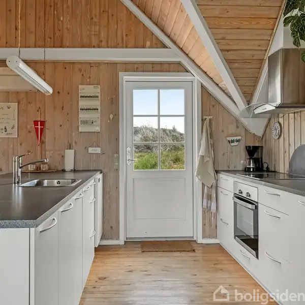Køkken med hvide skabe, stålgrej og indbygget ovn. Vasken er til venstre, og der er en dør til omgivelserne. Trævægge og loft med skråvinkel skaber en hyggelig atmosfære.