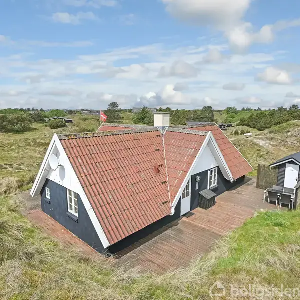 Et traditionelt sommerhus med rødt tegltag står på en træterrasse omgivet af en sandet naturgrund med græs og buske. En lille skurbygning ses ved siden af huset.
