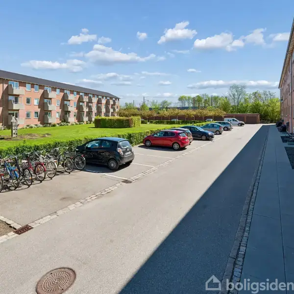 Parkeringsplads med biler holder stille langs cykler foran to røde murstensbygninger. Græsareal og buske omgrænser området med blå himmel og skyer i baggrunden.