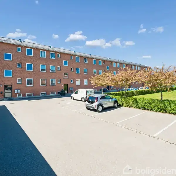 En lang, murstensbygning med mange vinduer står i et solrigt boligområde. To biler er parkeret på en asfaltvej foran bygningen. Blå himmel og træer omkranser stedet.