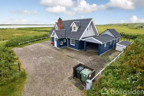 Et blåt hus med hvide kanter står på en grusbelagt gårdsplads, omgivet af grønt græsland. I baggrunden ses en sø og kuperet landskab under en blå himmel.