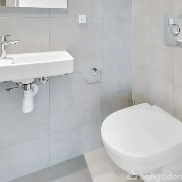 Håndvask med armatur monteret på væggen. I nærheden en væghængt toiletstol. Fliser i lyse nuancer dækker vægge og gulv. Minimalistisk design i et kompakt badeværelsesmiljø.