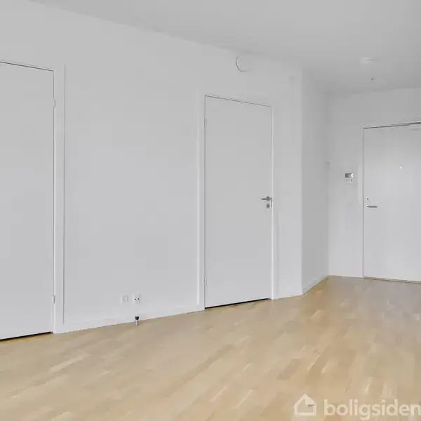 Døre står lukket i en lys gang med trægulve, omgivet af hvide vægge, og ingen tekst er synlig.