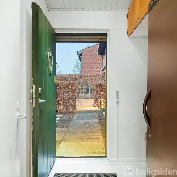 En åben grøn dør fører ud til en flisebelagt sti, omgivet af en lav hæk og et rødt murstenshus i baggrunden.