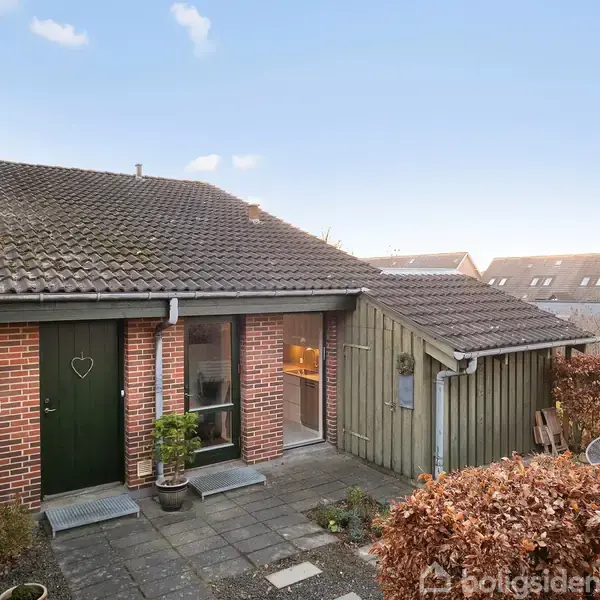 Et rødt murstenshus med sort tegltag har en grøn dør med et hjerte og store vinduer. Foran huset er der en flisebelagt gangsti omgivet af buske.