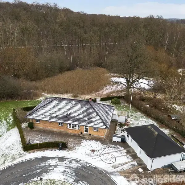 Et hus med et snedækket tag står ved siden af en garage i en skovklædt, sneklædt landskab. En rundkørsel omkranser ejendommen, mens skovstrækninger danner en baggrund.