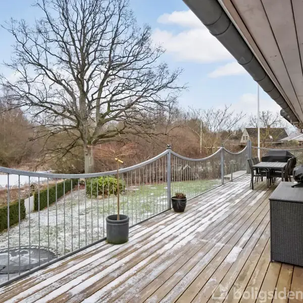 Træterrasse med en grill og møbler, omkranset af et gelænder, vender ud mod en stor have med en stor træ og vinterlandskab, himlen er blå med skyer.