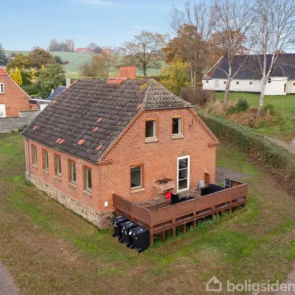 Det røde murstenshus står stille på en græsrabat omkranset af træer. Huset har et skråt tag og en træterrasse med affaldsbeholdere foran. Omkringliggende huse og veje ses i baggrunden.
