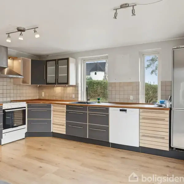 Køkken med sorte og lyse træskabe står ubenyttet i et rummeligt, lysfyldt rum. Komfur og opvaskemaskine er indbygget. Store vinduer giver udsigt til det grønne udenfor.