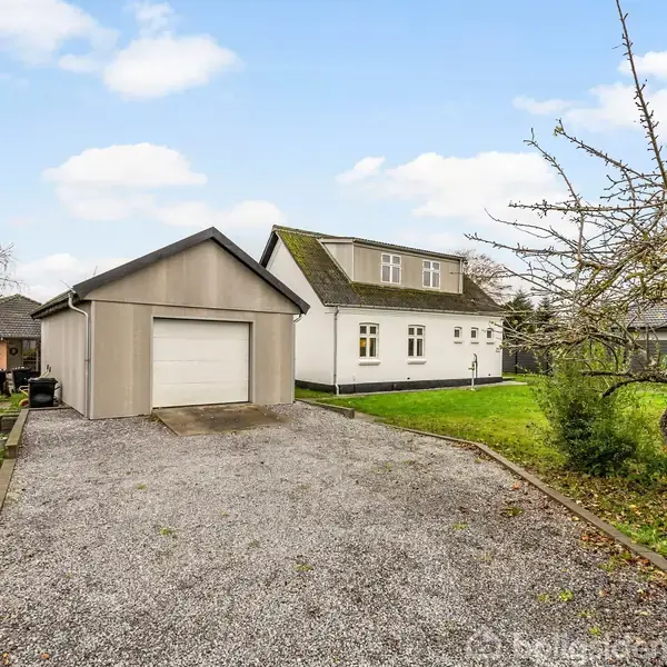 Et hvidt hus med en grusindkørsel og en garage ligger i en grøn have, omgivet af træer og buske, med en blå himmel og spredte skyer i baggrunden.