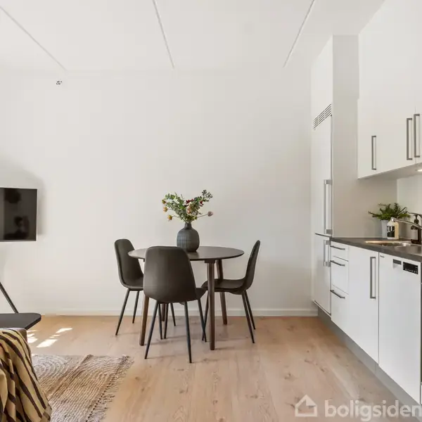 Et moderne køkken-alrum indeholder et rundt spisebord med fire stole og en vase med blomster. Til venstre står et tv på et stativ og en grøn plante.