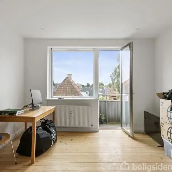 Et lyst rum med trægulv indeholder et skrivebord med computer ved siden af pakker og kasser. En åben dør fører til en balkon med udsigt over hustage og træer.