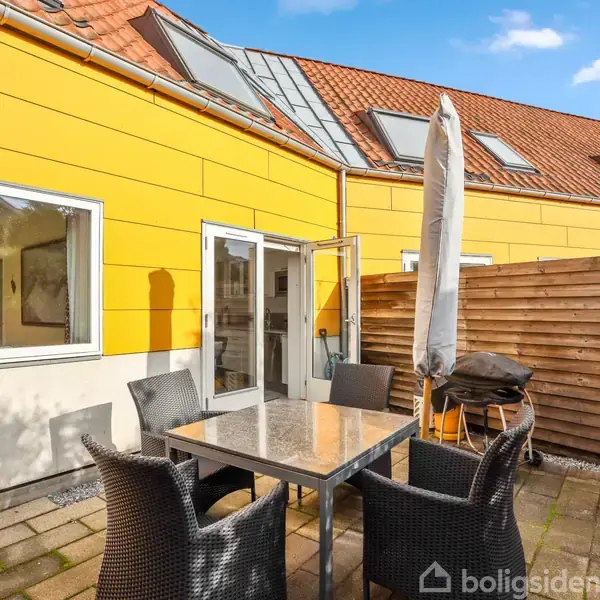 Udendørs terrasse med spisebord og fem stole. Parasol ved siden af bordet. Baggrund: gulmalet bygning med skråt tag og flere ovenlysvinduer. Flisebelagt gulv og træhegn til højre.