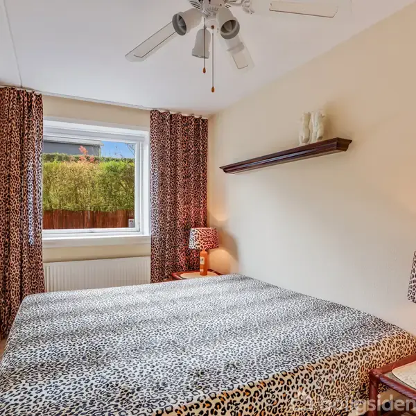 Soveværelse med stor sengebund med leopardmønstret sengetøj. En loftsventilator hænger over sengen. Der er et vindue med udsigt til en hæk, og der er lamper med matchende leopardmønster.