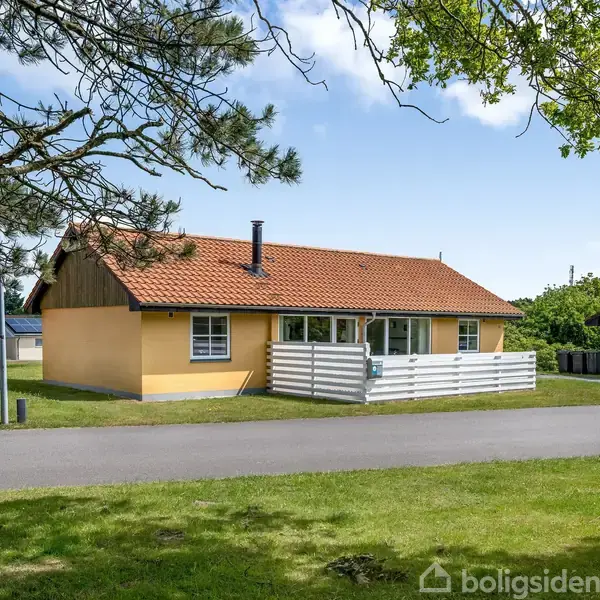 Et gult hus med rødt tegltag står stille ved en vej. Der er grønne træer og græsplæne omkring. En carport er delvist synlig til venstre.