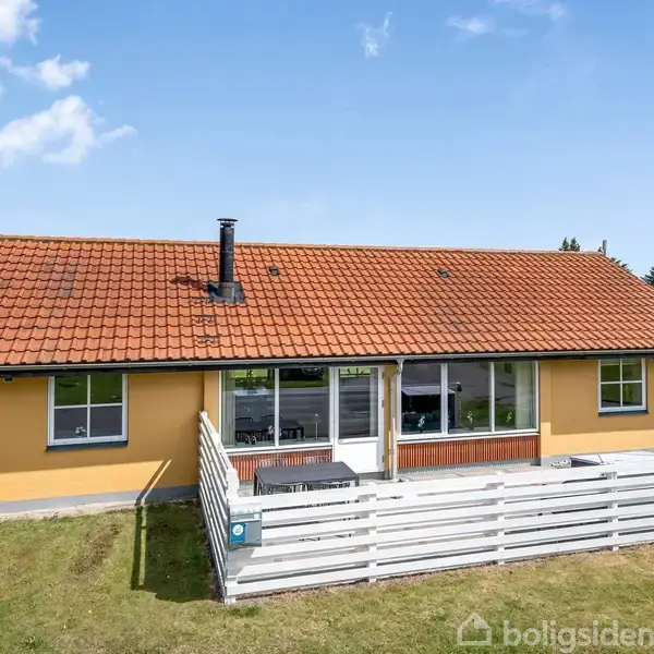 Et gult hus med rødt tegltag står stille i en grøn have. Foran er der en hvid træterrasse, omkranset af en lav hvid stakit.