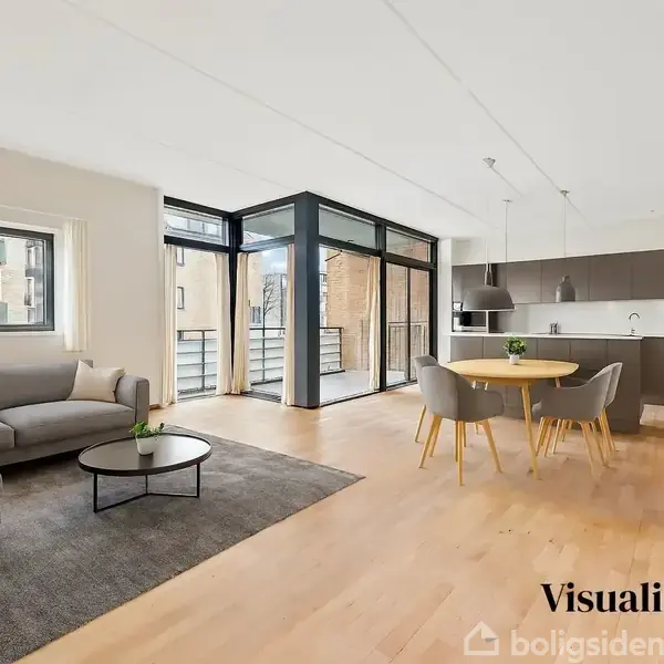 En moderne stue med en grå sofa ved siden af et rundt sofabord. Et spisebord med stole står ved et vindue. Køkkenet har indbyggede skabe. Tekst: "Visualisering".