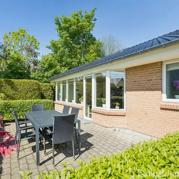 En rød murstensbygning med store vinduer vender ud mod en flisebelagt terrasse, hvor et sort bord med stole står. Omgivet af grønne hække og træer under en klar blå himmel.