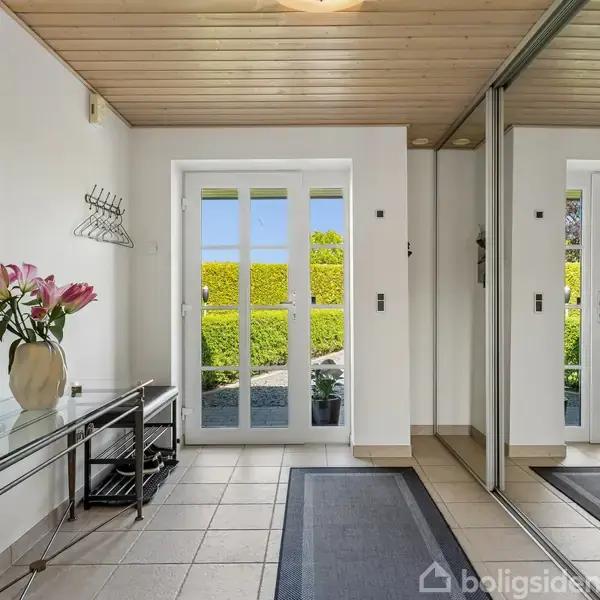 Entré med glasbord og vase med blomster. Dobbelt glasdør åbner ud til en have. Klinkegulv med tæppe, væg med spejl, træloft og knagerække til overtøj.
