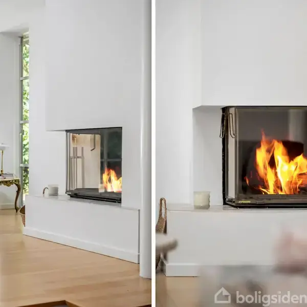 Pejs med brændende ild i en moderne stue med lyse trægulve. Venstre side viser også en grå sofa og et lille bord med lampe tæt ved store vinduer.