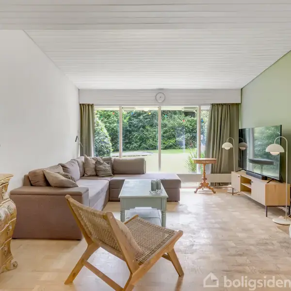 En stue med en stor, brun L-formet sofa står ved et glasbord. En kommode med en dekoreret lampe er ved siden af, og et fladskærms-tv er placeret overfor. Rummet har store vinduer, der åbner ud til en grøn have.