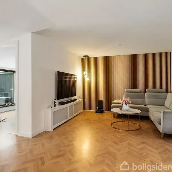 En stue med grå hjørnesofa står på trægulv, ved siden af et rundt sofabord. Til venstre hænger et TV på en lav reol, mens en væg med træpaneler oplyses af pendellamper.