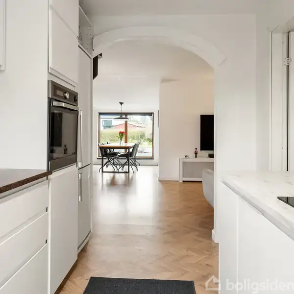 Overdækket terrasse med mørkt, fletsofaarrangement og kvadratisk bord beliggende på trædæk. Omgivet af højt hegn og buske. Åbent mod lyseblå himmel og omkringliggende huse.