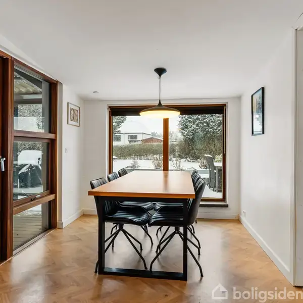 Spisebord med stole står i spisestue, oplyst af hængelampe. Store vinduer og dør giver udsigt til sneklædt have. Trægulv og hvidmalede vægge præger rummet.