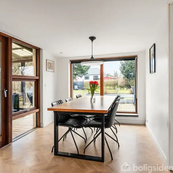 Et hvidt hus med mørkt tag ligger omkranset af en have med græsplæne. Huset har en terrasse med havemøbler og er omgivet af buske og træer i et villakvarter.
