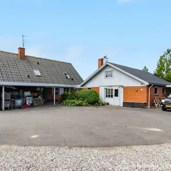 To huse med røde mursten står på en gårdsplads. En sort bil parkerer ved siden af, og omgivelserne inkluderer træer og en blå himmel.