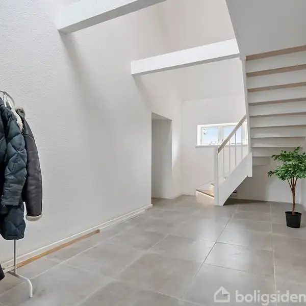 Tøjstativ holder jakker, stående ved væg; entré med gråt flisegulv. Hvid trappe fører opad. Indendørs plante i potte til højre. Minimalistisk og lys atmosfære.