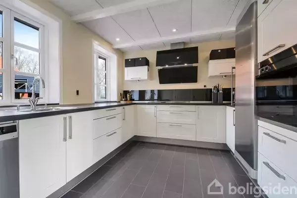 Et moderne køkken med hvide skabe og sorte bordplader. Ovn og køleskab i rustfrit stål står i højre side. Store vinduer slipper lys ind, omkranset af lyse vægge.