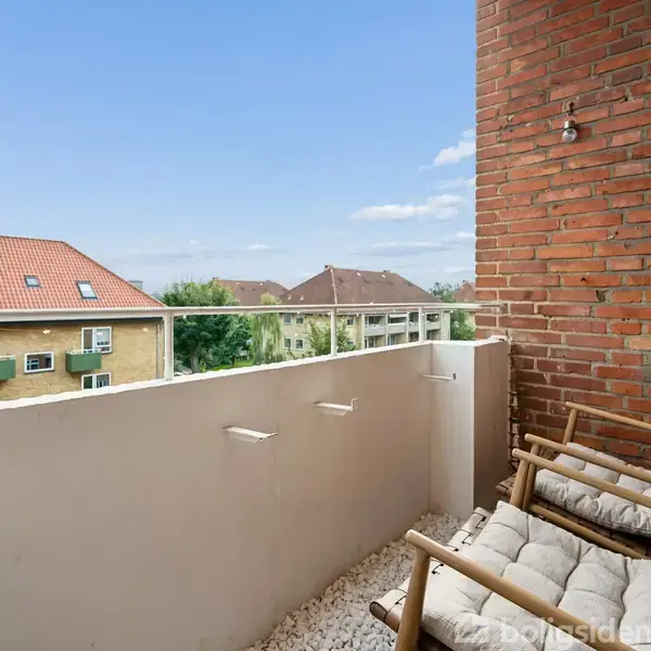 To loungestole med puder står på en lille altan med hvidt rækværk og udsigt til huse med røde tage. Murstensvæggene skaber en hyggelig atmosfære.