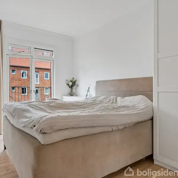 En dobbeltseng er placeret mod en væg med sengetøj let uordentlig. Rummet har hvidmalede vægge, et skab til højre og et vindue bagved, der viser en bygning udenfor.