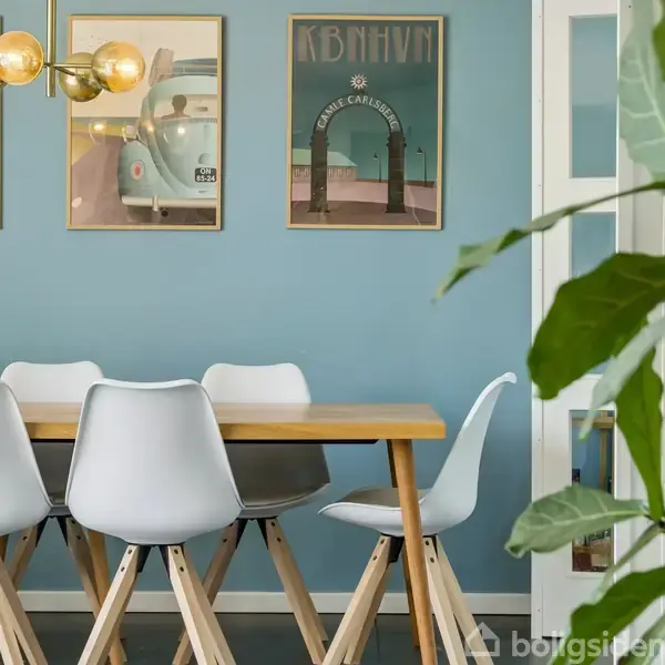Spisebord med hvide stole står i et rum med blå vægge. Over bordet hænger en moderne lampe. På væggen ses tre plakater med teksten "KBNHVN" og "DANISH CABINETMAKER".