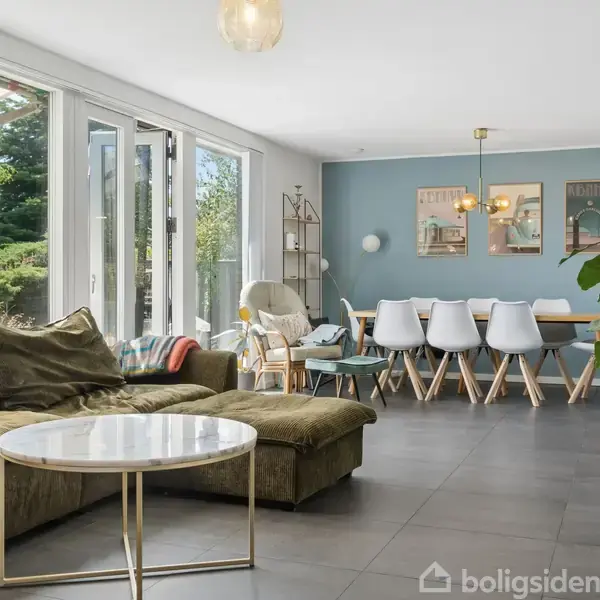 En stue med en stor grøn sofa og rundt, hvidt sofabord. Et spisebord med hvide stole står i baggrunden. Rummet har store vinduer, som fører ud til en have.