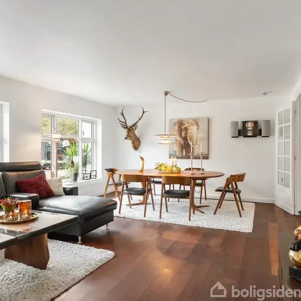 En stue med en grå hjørnesofa og et sofabord dekoreret med lys og pyntegenstande. I baggrunden står et spisebord med stole. Lyse vægge og parketgulv omkranser rummet.