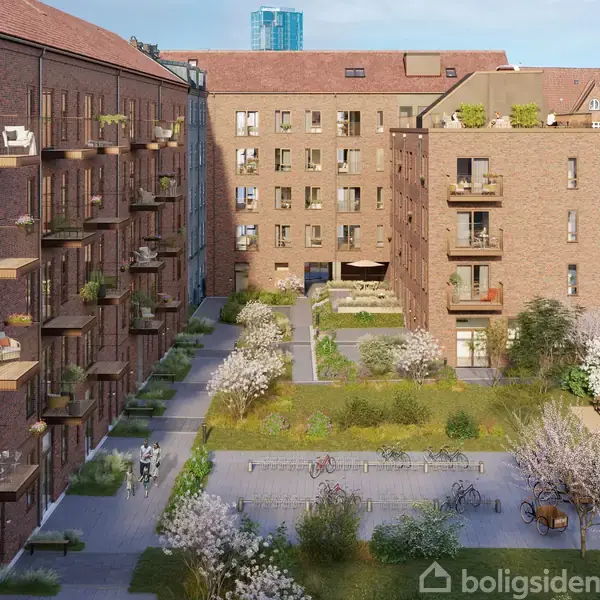 Bygninger i mursten omkranser en gårdhave med frodig grøn beplantning og træer. Altaner strækker sig ud fra facaderne, og cykelstativer står ved siden af stier og græsarealer.
