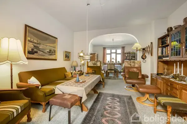 Stue med brun sofa og lænestole omkring et sofabord med dug. Rummelige reoler fyldt med bøger står langs væggene. En bred døråbning fører til et tilstødende spisestueområde.