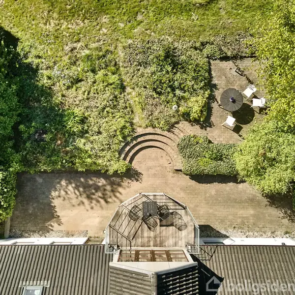 Tagterrasse med udsigt over en frodig have. Havemøbler står ved et rund havebord med parasol i et aflukket hjørne af haven, omgivet af buske og træer.